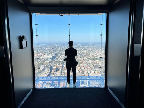 A person stands in a glass-walled observation area, overlooking a city skyline from a high vantage point.