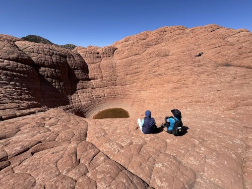A person sits on rocky terrain near a shallow pool, surrounded by textured red rock formations under a clear blue sky.