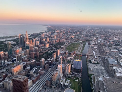 Aerial view of a city skyline at sunset, featuring buildings, a river, and the coastline in the distance.