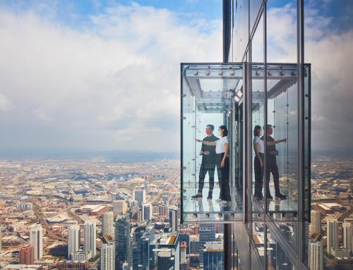 Two people in a glass elevator overlooking a city skyline, with clouds and blue sky in the background.