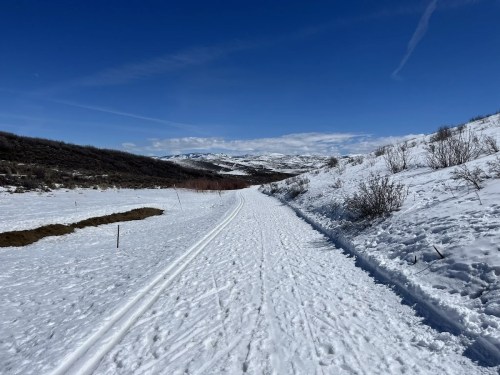 A snowy trail winds through a winter landscape, surrounded by hills and a clear blue sky.