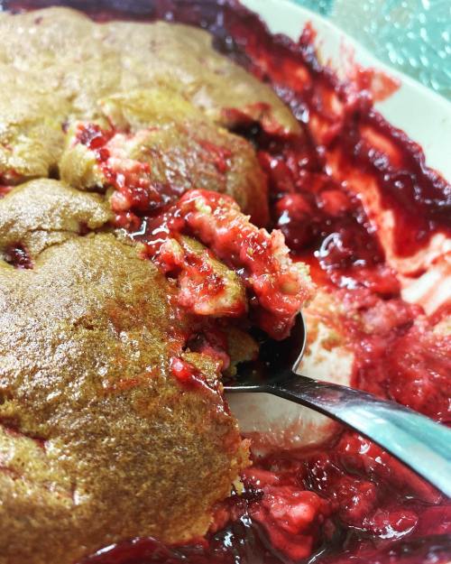 A close-up of a spoon digging into a warm berry dessert with a golden-brown topping and vibrant red filling.