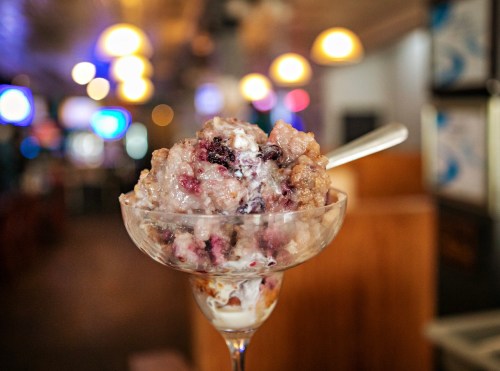 A close-up of a dessert in a glass, featuring layers of ice cream and mixed berries, with a blurred background.