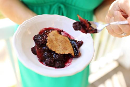 A person holds a bowl of berry cobbler with a fork, featuring a golden crust and dark berry filling.