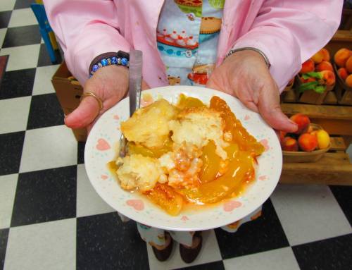 A person holds a plate of peach cobbler topped with syrup, surrounded by a checkered floor and fruit baskets.