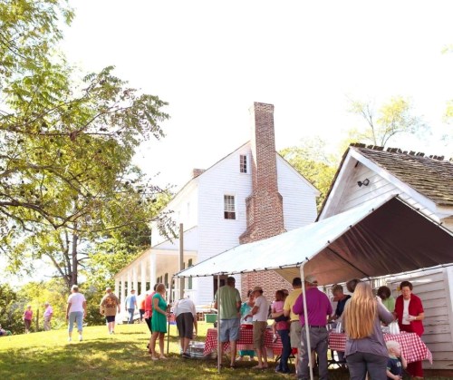 A gathering of people at an outdoor event near a white house, with tables under a tent and trees in the background.
