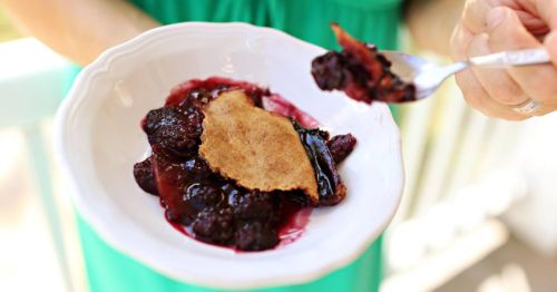 A person holds a fork with a piece of berry cobbler above a bowl filled with mixed berries and a golden crust.