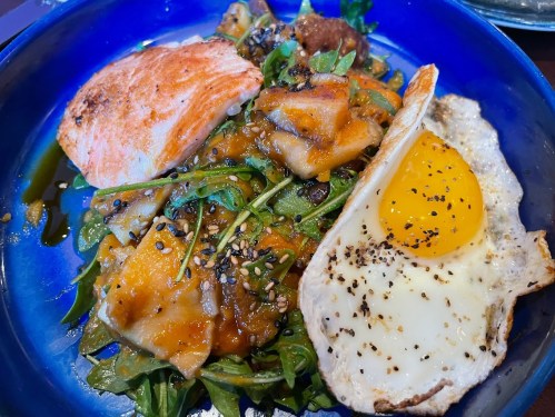 A vibrant blue bowl filled with salmon, arugula, sweet potatoes, and a sunny-side-up egg, garnished with sesame seeds.