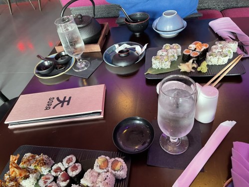 A table set with sushi, a teapot, drinks, and a menu at a restaurant.