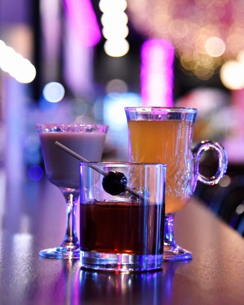 Three colorful cocktails on a bar, featuring a cherry garnish, with a blurred, vibrant background.