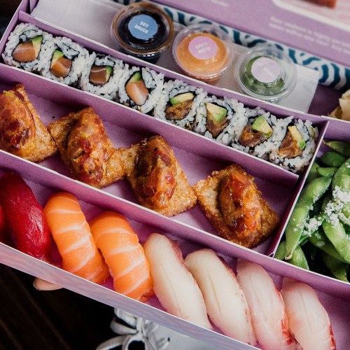 A colorful sushi platter featuring rolls, sashimi, and edamame in a pink box.
