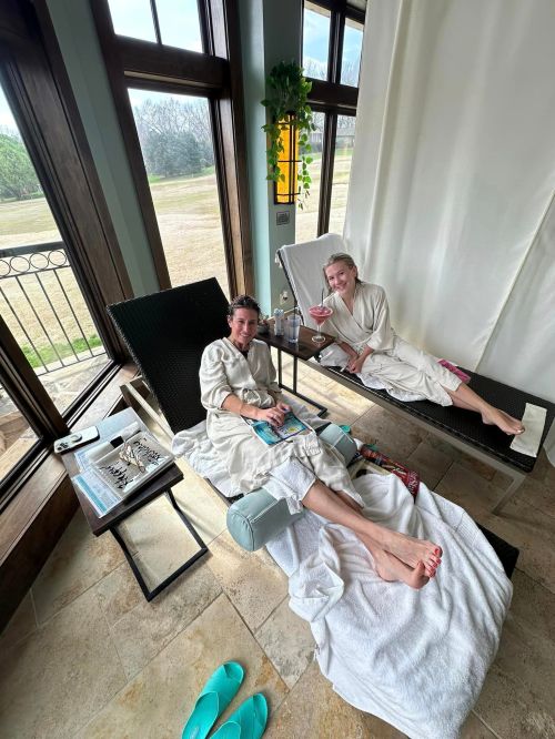 Two women in bathrobes relax on lounge chairs, enjoying drinks and a spa day in a bright, airy room with large windows.