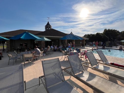 A sunny poolside scene with lounge chairs, umbrellas, and people socializing near the water.