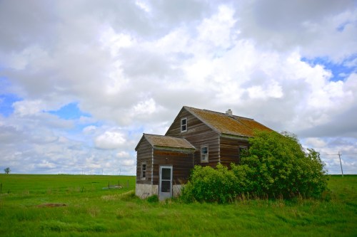 A weathered wooden house surrounded by green fields under a cloudy sky.