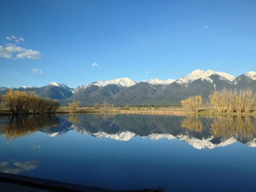 A serene lake reflecting snow-capped mountains and clear blue skies, with trees lining the shore.