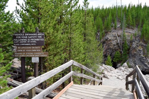 A wooden walkway leads to a swimming area, with a sign listing safety rules and prohibitions in a forested setting.