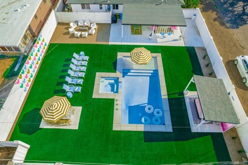 Aerial view of a backyard with a pool, lounge chairs, umbrellas, and a shaded seating area on green turf.