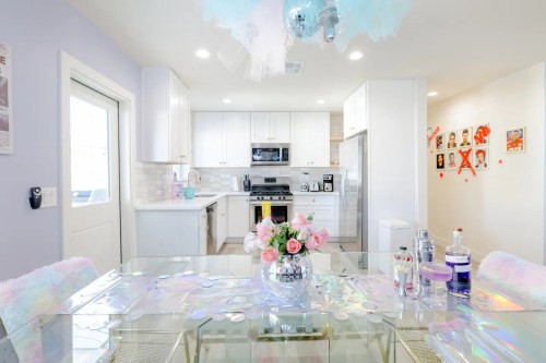 Bright kitchen and dining area with a glass table, pastel decor, and a floral centerpiece. Modern appliances in the background.