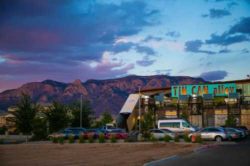 A colorful building with "Tim Can Alley" sign, surrounded by cars and trees, with mountains and a sunset in the background.