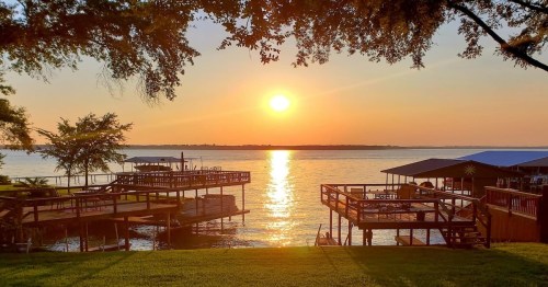 a lakeside view of two wooden decks with lots of chairs and trees above in front of a golden sunset by the water