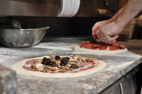 A hand sprinkles seasoning on a pizza topped with pepperoni, while another pizza with mushrooms sits nearby on a floured counter.