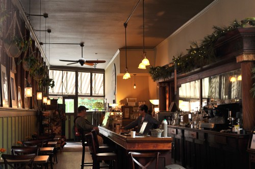 A cozy café interior with wooden furniture, a bar counter, and warm lighting, featuring two people engaged in conversation.