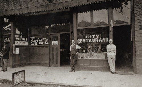 Historic storefront featuring the Carbon Hotel and City Restaurant, with two men standing outside. Black and white photo.