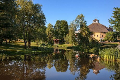 A serene landscape featuring a pond reflecting trees and a round building under a clear blue sky.