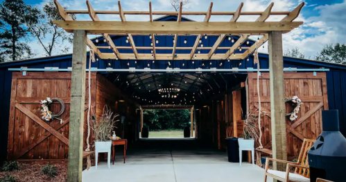 A rustic barn entrance with wooden doors, string lights, and decorative wreaths, set against a blue sky.