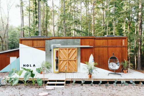 A modern cabin with a wooden exterior and a large deck, surrounded by trees and greenery.