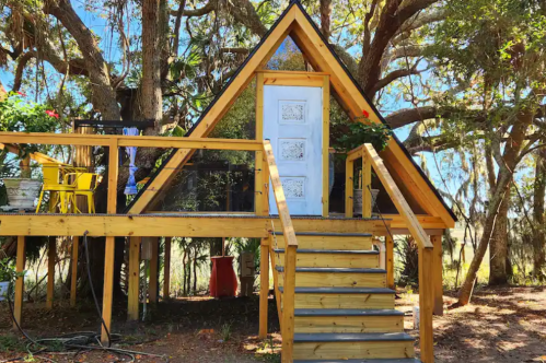 A charming wooden A-frame cabin nestled among trees, featuring a white door and a staircase leading up to it.