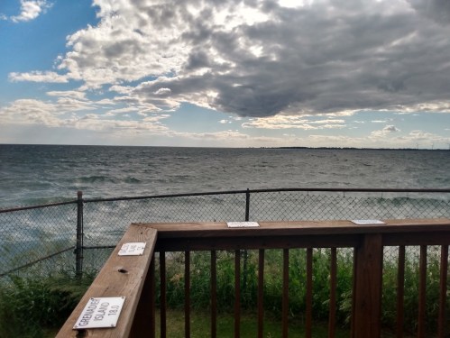 A view of a lake under a cloudy sky, with a wooden railing and signs in the foreground.