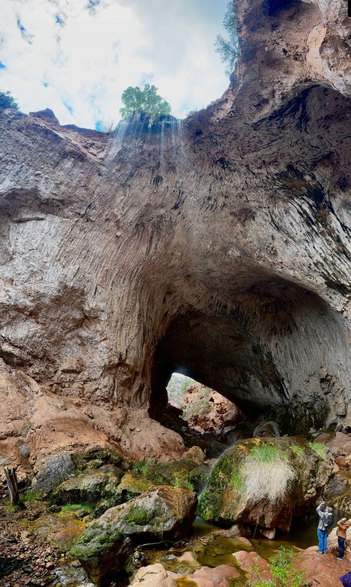 A large rock arch with a waterfall, surrounded by lush greenery and rocky terrain, under a cloudy sky.