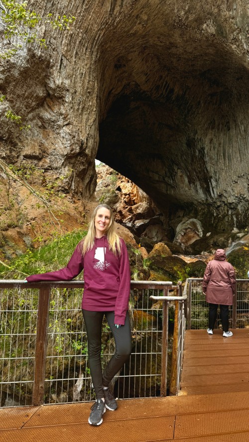 A woman in a maroon sweatshirt stands by a railing in a cave, with a person in the background exploring the area.