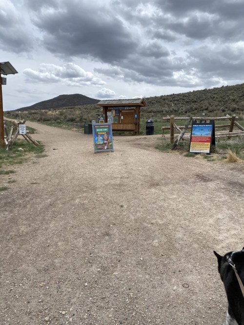 A dirt path leads to a visitor center with signs, surrounded by grassy hills and cloudy skies.