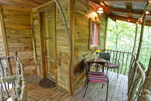 A cozy wooden cabin entrance with a small table and chairs on a balcony surrounded by trees.