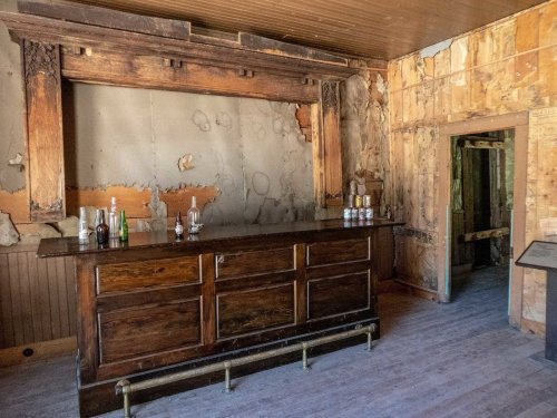 A rustic, weathered bar with bottles on top, set in a dilapidated room with peeling walls and wooden accents.