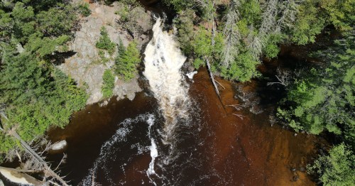Aerial view of a waterfall cascading into a dark river, surrounded by lush green trees and rocky terrain.