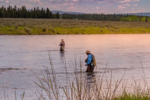 Two anglers wade in a river, surrounded by lush greenery and distant trees under a colorful sky.