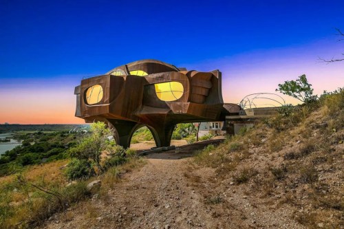 A unique, modern building with a rust-colored exterior, elevated on stilts, surrounded by greenery and a sunset sky.