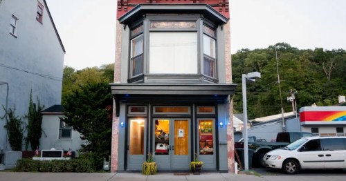 Symmetrical two-story historic building with a grey facade, red roof accents, and a large second-floor bay window.