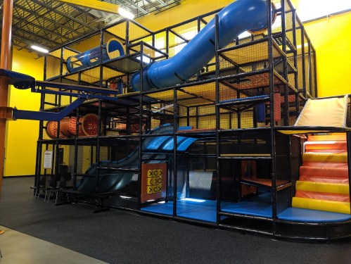 Colorful indoor play structure with slides, tunnels, and climbing areas against a bright yellow wall.