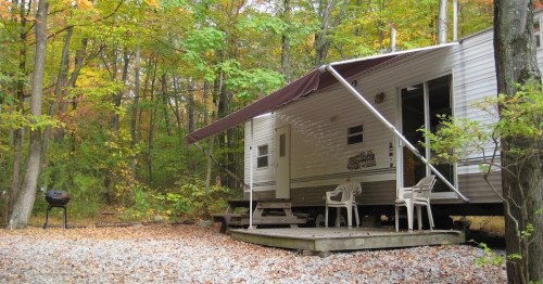 A cozy trailer in a wooded area, surrounded by autumn leaves, with a porch and outdoor seating.