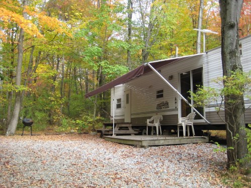 A cozy trailer in a forested area with autumn leaves, featuring a porch, chairs, and a grill nearby.