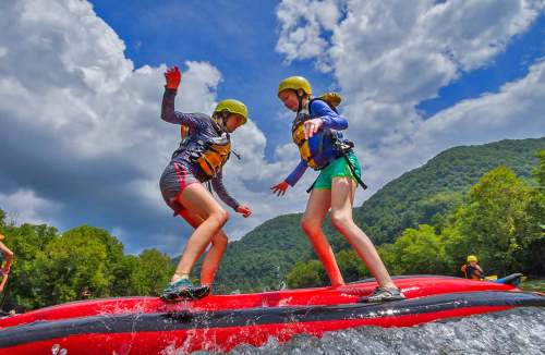 Two girls in helmets balance on a red inflatable raft while whitewater rafting, surrounded by green hills and a blue sky.