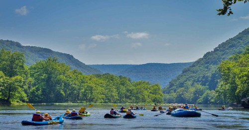 A group of people kayaking on a calm river surrounded by lush green hills under a clear blue sky.