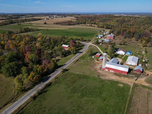 Aerial view of a rural landscape featuring farms, a silo, and colorful autumn trees along a winding road.