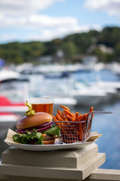 A burger with lettuce and onion, sweet potato fries in a basket, and a glass of beer, with a marina in the background.