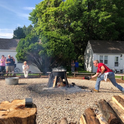 A man in a red shirt tends to a fire pit while onlookers watch from a distance in a sunny outdoor setting.