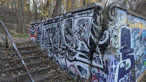 A weathered concrete wall covered in colorful graffiti, surrounded by autumn foliage and a set of worn stairs.